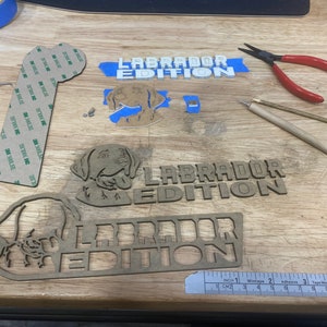 May include: Three wooden cutouts of a Labrador Retriever dog with the text "Labrador Edition" on each. The cutouts are on a wooden surface with a ruler, tape, and other tools.