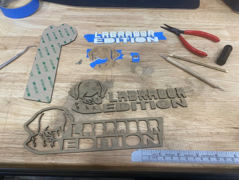 May include: Three wooden cutouts of a Labrador Retriever dog with the text "Labrador Edition" on each. The cutouts are on a wooden surface with a ruler, tape, and other tools.