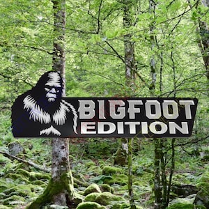 May include: A black sign with the words "BIGFOOT EDITION" in silver lettering, featuring a silhouette of Bigfoot. The sign is set against a backdrop of a lush green forest with moss-covered ground.