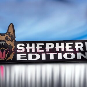 以下が含まれることがあります： ジャーマンシェパードの犬の頭と「SHEPHERD EDITION」の文字が書かれた黒と銀のエンブレムです。
