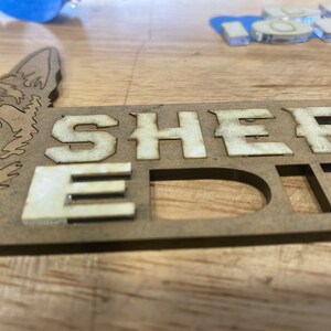 以下が含まれることがあります： 白い文字で「SHEPHERD EDIT」と書かれた木製の看板。看板は犬の頭の切り抜きで飾られています。