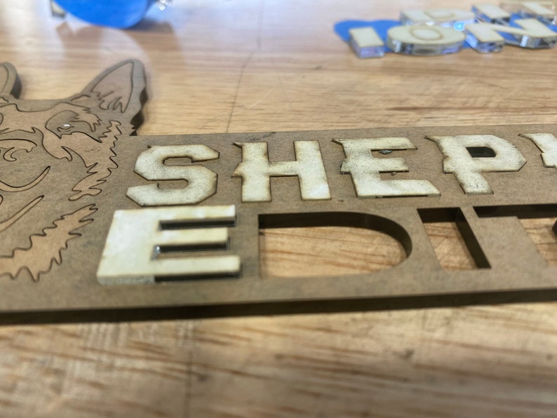 以下が含まれることがあります： 白い文字で「SHEPHERD EDIT」と書かれた木製の看板。看板は犬の頭の切り抜きで飾られています。