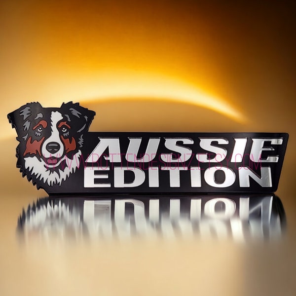 May include: Aussie Edition emblem featuring an Australian Shepherd dog head. The emblem has a black background with silver lettering and a detailed dog illustration in black, white, and brown. The image has a reflective surface.