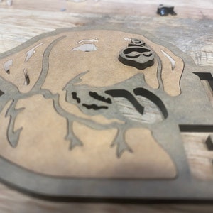 May include: A wooden cutout of a dog's head, with a brown stain finish. The dog's head is facing to the right, and the cutout is detailed with the dog's ears, nose, and mouth.