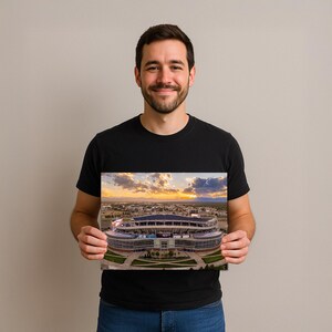 A man is holding a framed photograph of a stadium, smiling and looking directly at the camera.