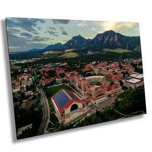 May include: An aerial view of a university campus with red-roofed buildings, a stadium, and a large mountain range in the background. The sky is cloudy with a hint of sunlight. The campus is surrounded by green trees and a road.