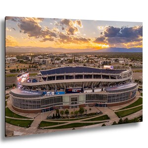 An aerial view of a large sports stadium with a curved roof and multiple levels, set against a backdrop of a colorful sunset sky.