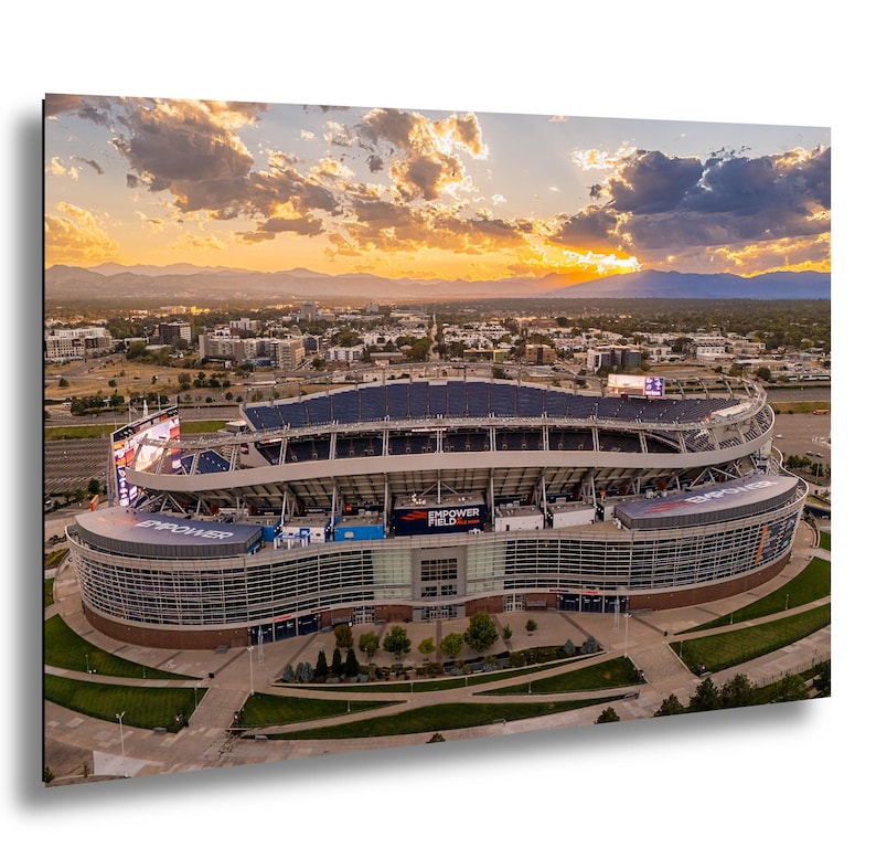 An aerial view of a large sports stadium with a curved roof and multiple levels, set against a backdrop of a colorful sunset sky.