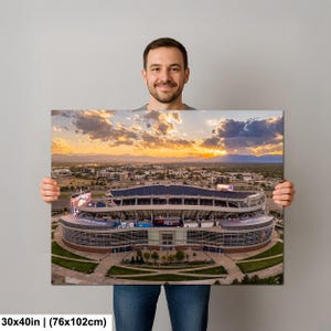 A man is holding a large framed print of an aerial view of a stadium, with a beautiful sunset in the background.