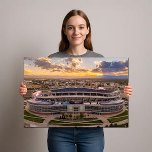 A woman is holding a framed photograph of a large stadium with a beautiful sunset in the background.