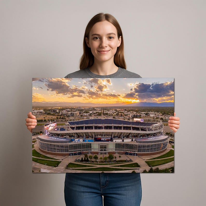 A woman is holding a framed photograph of a large stadium with a beautiful sunset in the background.