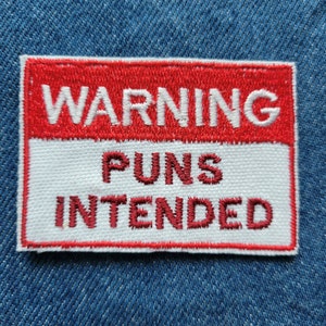 Puede incluir: Un parche bordado rojo y blanco con el texto "WARNING PUNS INTENDED" en letras blancas sobre fondo rojo y letras rojas sobre fondo blanco.