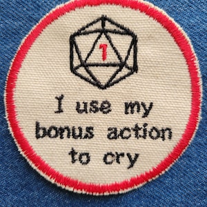 May include: A white circular patch with a red border. The patch features a black and white illustration of a 20-sided die with a red "1" on it. The text "I use my bonus action to cry" is embroidered in black below the die.