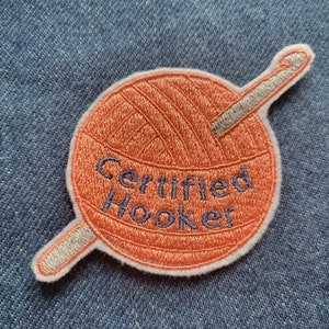 May include: A round orange yarn ball with a silver crochet hook sticking out of it. The yarn ball has the words "Certified Hooker" embroidered on it in blue thread.