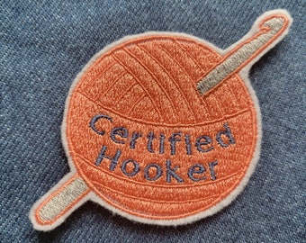Puede incluir: Una bola de hilo naranja redonda con un gancho de ganchillo plateado que sobresale. La bola de hilo tiene las palabras "Certified Hooker" bordadas en hilo azul.