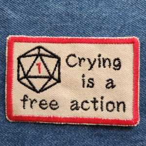 Puede incluir: Un parche de tela blanco con un borde rojo. El parche presenta una ilustración en blanco y negro de un dado de 20 caras con un "1" rojo en él. El texto "Crying is a free action" está bordado en negro en el parche.