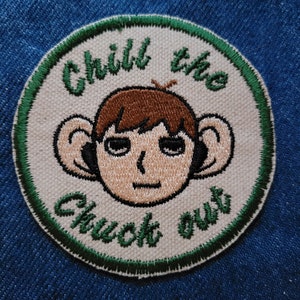 May include: A white embroidered patch with a green border. The patch features a cartoon illustration of a person with brown hair and large ears. The text "Chill the chuck out" is embroidered in green around the person's head.