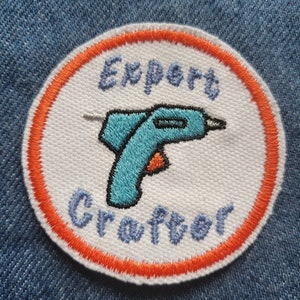 May include: A white round embroidered patch with an orange border. The patch features a blue and black cartoon glue gun and the text "Expert Crafter".