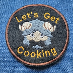 May include: A black and brown embroidered patch featuring a blue and white cartoon character with horns. The text "Let's Get Cooking" is embroidered in yellow around the character.