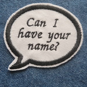 Op de afbeelding: Witte geborduurde patch met zwarte stiksels in de vorm van een spreekballon. De tekst in de ballon luidt "Can I have your name?"