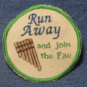 Puede incluir: Un parche redondo con ribete verde con el texto "Run Away and Join the Fa" bordado en verde y azul. Un instrumento de viento de bambú bordado en marrón se representa debajo del texto.