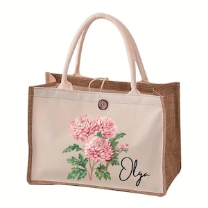 Puede incluir: Un bolso tote rectangular de yute con un panel frontal de lona color crema. El panel presenta un diseño floral de crisantemos rosas y hojas verdes, con el nombre "Olga" en escritura negra. El bolso tiene asas color crema y un cierre de botón de madera.