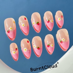 May include: Ten pink and white false nails with a strawberry and flower design. The nails are arranged in two rows on a blue surface.
