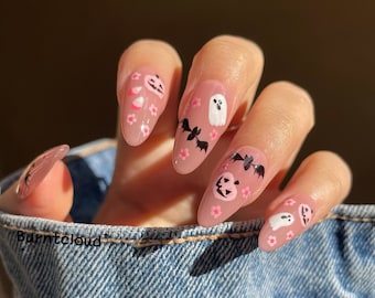 May include: Pink almond-shaped nails with Halloween-themed nail art. The designs include ghosts, bats, and pumpkins with pink flowers.