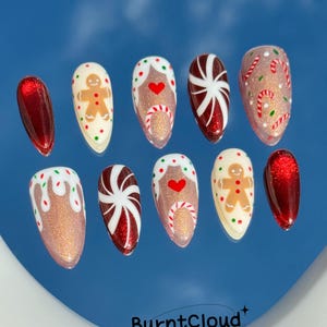May include: A set of ten festive, almond-shaped press-on nails. Designs include gingerbread men, candy canes, and red and white peppermint swirls, all set against a glittery, nude base. The nails are arranged on a reflective blue surface.