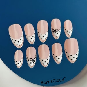 May include: A set of ten oval false nails. Each nail has a light pink base with a white tip decorated with black polka dots. Two nails feature a black bow design. The brand name "BurntCloud" is visible.