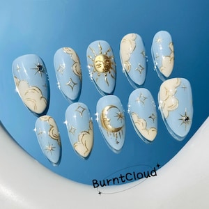 May include: A set of ten light blue press-on nails with gold celestial designs. Each nail features a different design, including clouds, stars, a sun, and a moon. The brand name "BurntCloud" is printed on the bottom right.