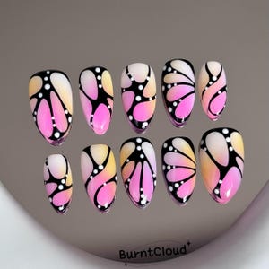 Könnte beinhalten: Ein Set aus zehn künstlichen Fingernägeln mit einem Farbverlauf von sanftem Gelb zu leuchtendem Pink, umrandet von kräftigen schwarzen Linien und akzentuiert mit kleinen weißen Punkten. Die Nägel haben eine glatte, glänzende Oberfläche und sind in zwei Reihen angeordnet.
