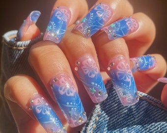 May include: Close-up of a hand with long, blue and pink ombre press-on nails. The nails have a floral design and glitter accents.