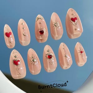 May include: Set of ten almond-shaped false nails with a light pink base and gold and red accents. Each nail features a different design with stars, moons, hearts, and sun symbols.