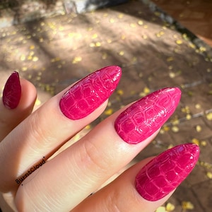 Puede incluir: Primer plano de una mano con uñas largas en forma de almendra pintadas de magenta vibrante. Las uñas tienen un efecto texturizado de piel de cocodrilo. El nombre de la marca "BurntCloud" es visible en la mano.