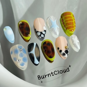 May include: A set of ten false nails with various designs. The designs include a blue and white polka dot, a brown and white animal print, a brown and green striped design, a white star, a brown and white cow print, and a brown and white leopard print. The nails are arranged on a white surface with the text "BurntCloud" in black.