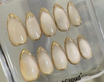May include: A set of ten false nails with a light beige base and gold accents. The nails are arranged in a clear plastic container.