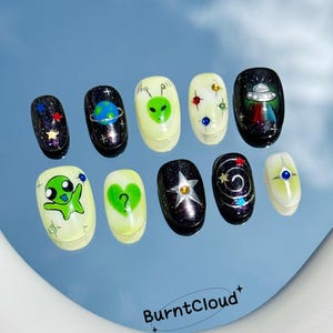 May include: A set of ten press-on nails with an alien theme. Designs include planets, stars, and alien faces in green, black, and white. Some nails have glitter accents. The brand name "BurntCloud" is at the bottom.