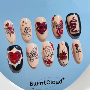 May include: Set of ten artificial nails with various designs, including hearts, butterflies, roses, and a snake. The nails are painted in a nude color with black, red, and white accents. The brand name "BurntCloud" is at the bottom.