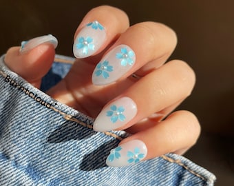 May include: White almond-shaped nails with blue flower accents. The nails are painted white and have a subtle shimmer. Each nail features a blue flower with a pearl center.
