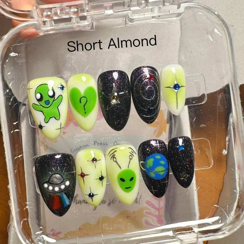 K&ouml;nnte beinhalten: Ein Set kurzer, mandelf&ouml;rmiger Press-On-N&auml;gel mit verschiedenen Alien-Designs. Die N&auml;gel weisen eine Mischung aus Farben auf, darunter Gr&uuml;n, Schwarz und Gelb, mit Glitzer und Cartoon-Illustrationen. Der Text "Short Almond" ist sichtbar.