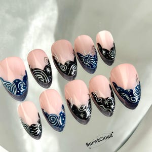 May include: A set of ten almond-shaped false nails. Each nail features a light pink base with a dark blue and black design at the tip. The designs include celestial elements like the sun, moon, stars, and swirling patterns. The brand name "BurntCloud" is visible.