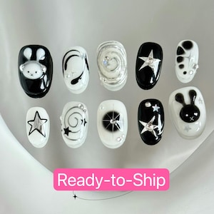 May include: A set of ten press-on nails in various designs. The nails feature black and white colors with star, swirl, and bunny designs. Some nails have silver embellishments. The text "Ready-to-Ship" is displayed on a pink rectangle.