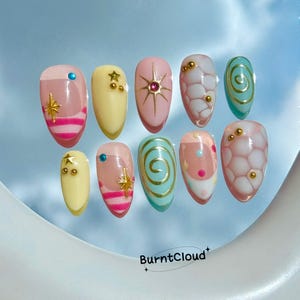 May include: A set of ten press-on nails featuring various designs including pink and yellow bases, gold star accents, blue and pink dots, white honeycomb patterns, and gold swirls. The text BurntCloud is printed at the bottom of the image.