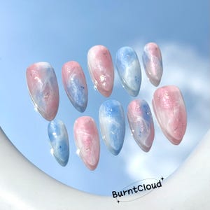 Peut inclure: Un ensemble de dix faux ongles en forme d'amande présentant un motif marbré dans des tons de rose pastel, bleu ciel et blanc avec de subtils reflets pailletés. Le texte BurntCloud est imprimé en noir dans le coin inférieur droit de l'image.