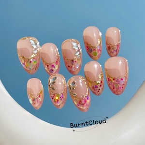 May include: Set of ten artificial nails with a light pink base, gold trim, and colorful glitter accents. Some nails feature silver leaf-shaped embellishments. The nails are oval-shaped and arranged on a reflective blue surface.