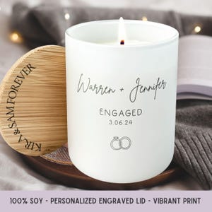 May include: A white candle with a lit wick, featuring the names "Warren + Jennifer", the word "ENGAGED", and the date "3.06.24". The candle has a wooden lid engraved with "KIRA & SAM FOREVER". The candle is on a wooden tray.