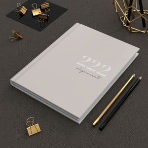 May include: A white journal with the number 222 and the word "alignment" written on the cover. The journal is on a gray surface with two pencils and gold paper clips.