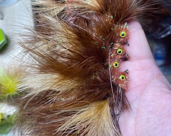 May include: A close-up of a handmade fishing fly. The fly features brown and tan feathers, with small, orange-brown bodies and yellow eyes. Iridescent accents are visible. The fly is designed to imitate an insect.