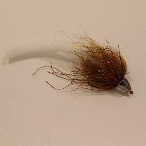 May include: A handmade fishing fly with a silver head and a body of brown and tan fibers. The fly has long, dark brown strands extending from the body and a white, fluffy tail. This fly is designed for catching fish.